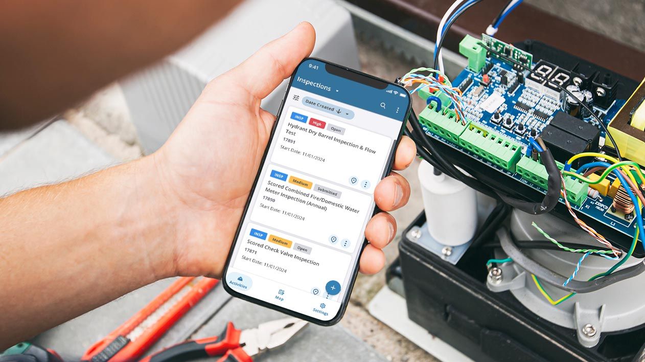 A technician holds a smartphone displaying a list of inspections, while working on an open electrical box with visible wires and a circuit board in the background.