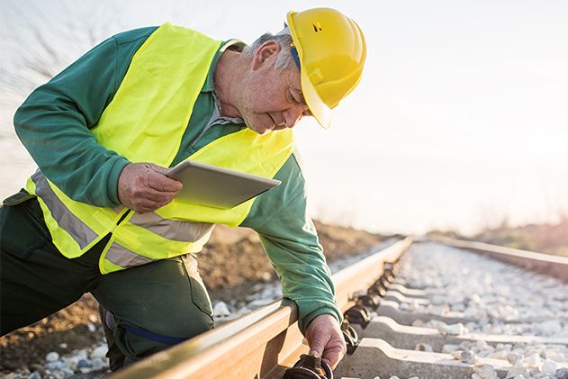 Ein Arbeiter mit gelbem Schutzhelm und Weste inspiziert ein Bahngleis und hält dabei ein Tablet in den Händen.