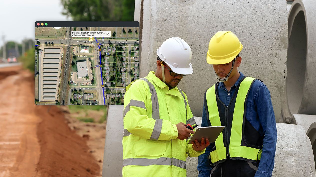 Deux ouvriers du bâtiment, portant un casque et un gilet de sécurité, regardent une carte numérique sur une tablette montrant une route et une ligne bleue représentant un service public.