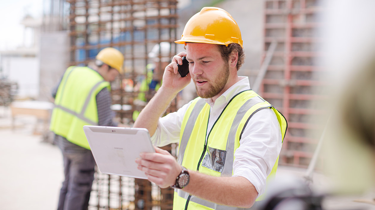 Ein Mann auf einer Baustelle mit gelbem Schutzhelm und Sicherheitsweste telefoniert und hält dabei ein Tablet in den Händen.