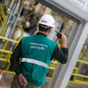 Ein Arbeiter in grüner Weste und weißem Schutzhelm mit der Aufschrift „OPERAÇÃO“ steht in einer Industrieanlage mit gelben Geländern und Maschinen und kommuniziert über Funk.