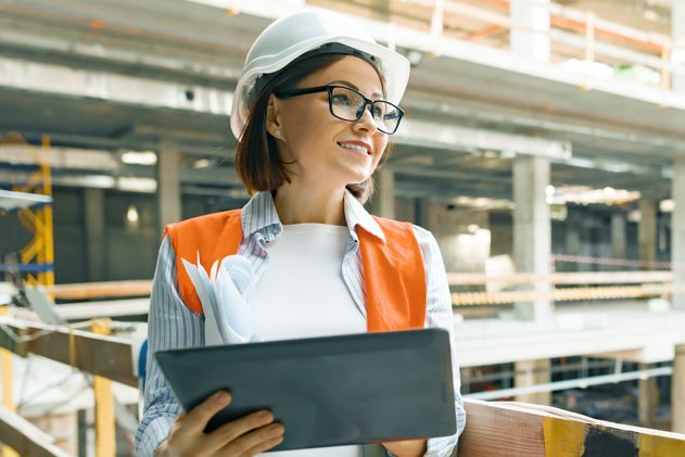 Eine lächelnde Frau mit Schutzhelm und orangefarbener Weste hält auf einer Baustelle ein Tablet und Papiere in der Hand.