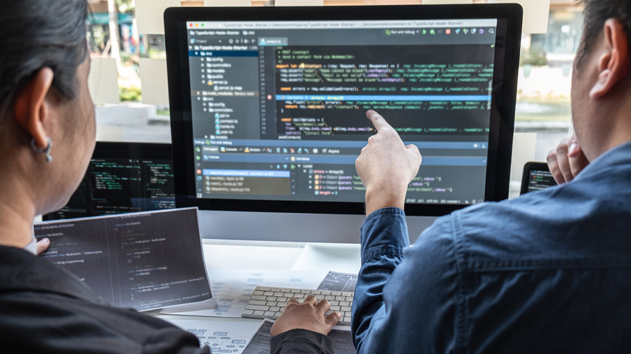 Zwei Personen arbeiten an einem Schreibtisch auf einem großen Monitor, auf dem komplexer Code angezeigt wird, wobei eine Person auf den Bildschirm zeigt.