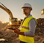A construction worker in a safety vest uses a Trimble rugged tablet at a job site with a yellow excavator in the background.