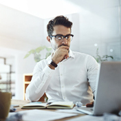 Ein Mann mit Brille sitzt an einem Schreibtisch mit einem Laptop und denkt nach.