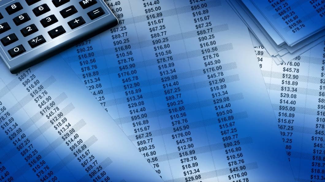 A calculator rests on a stack of papers filled with rows of dollar amounts, with a blue hue.