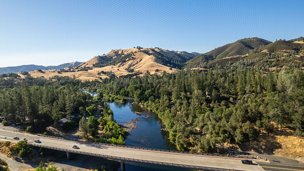 Aerial view of a multi-lane concrete highway bridge carrying vehicles over a river, surrounded by golden, tree-covered hills under a clear sky.