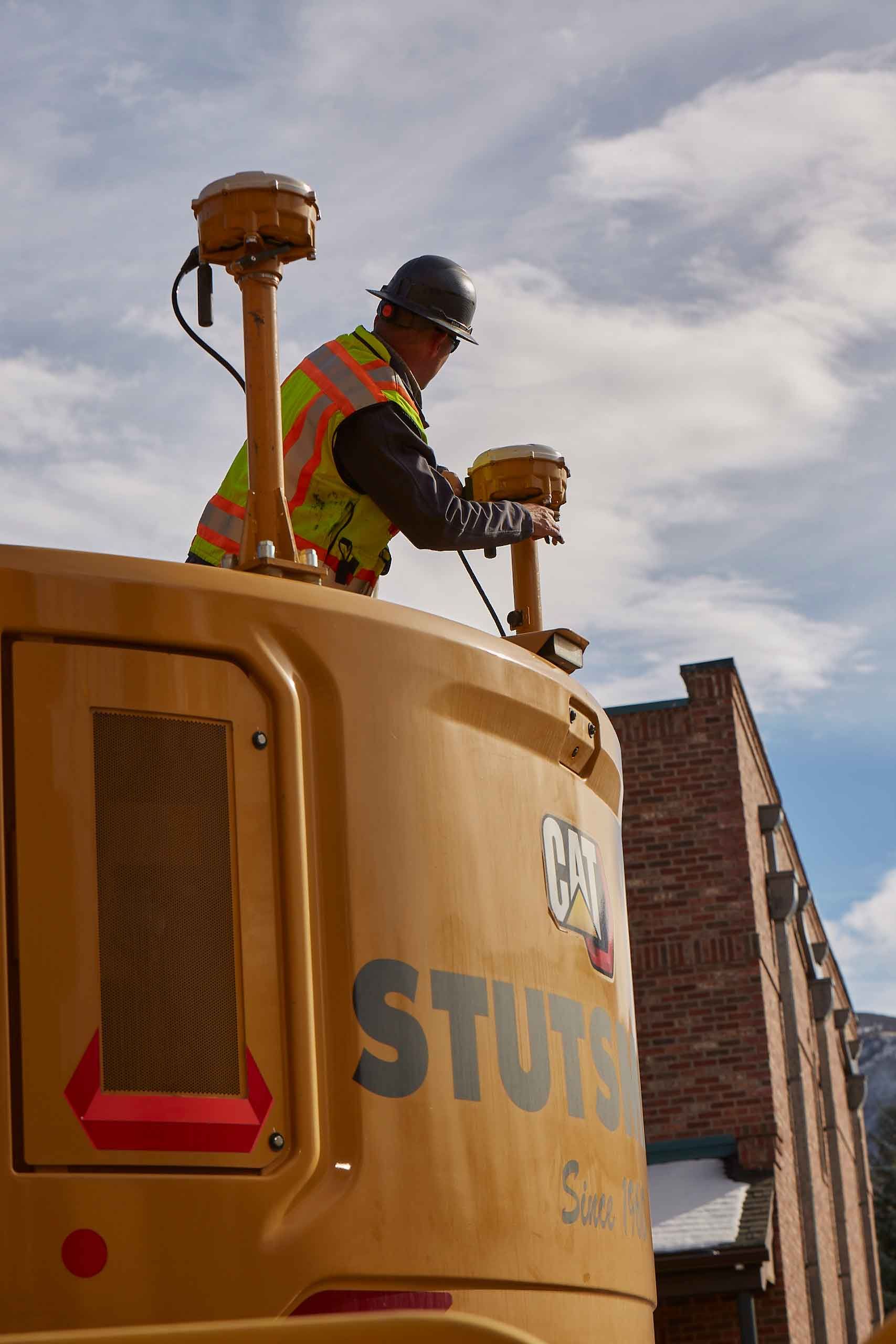 A worker in a hard hat and safety vest adjusts a GPS receiver on a Caterpillar machine.