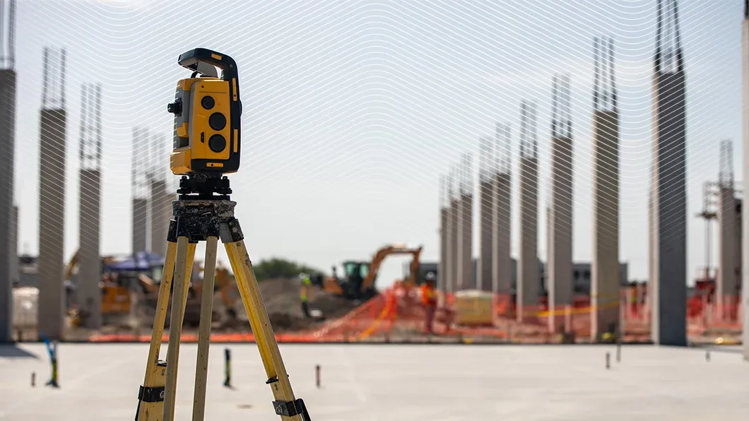A yellow Trimble 3D laser scanner on a tripod at a construction site with concrete pillars and an excavator in the background.