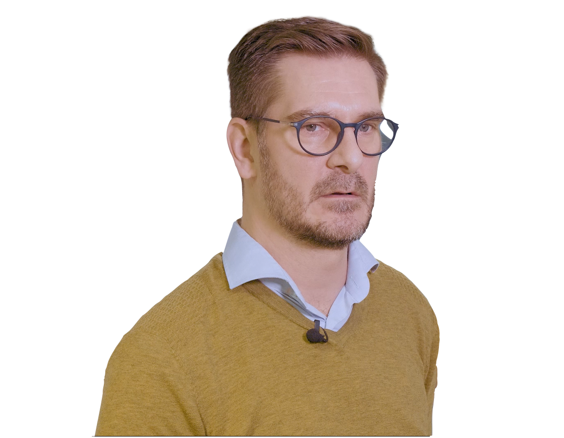 A man with short brown hair, glasses, a light blue collared shirt, and a mustard yellow sweater is shown from the chest up.