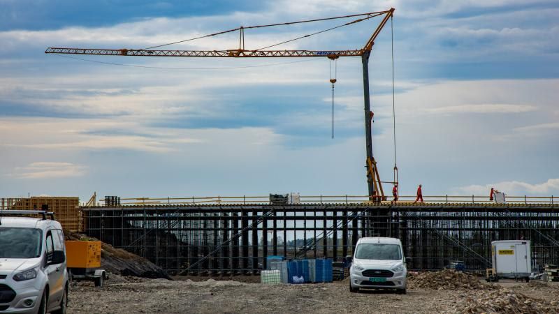 Construction site with a large crane and workers on a bridge under construction, with vans and equipment in the foreground.