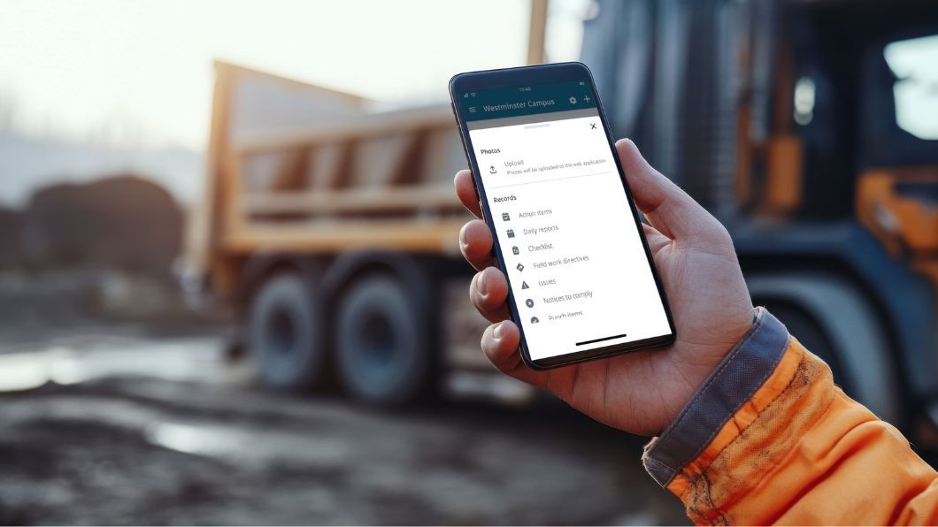 A worker holds a smartphone displaying a the ProjectSight mobile app on an outdoor site.