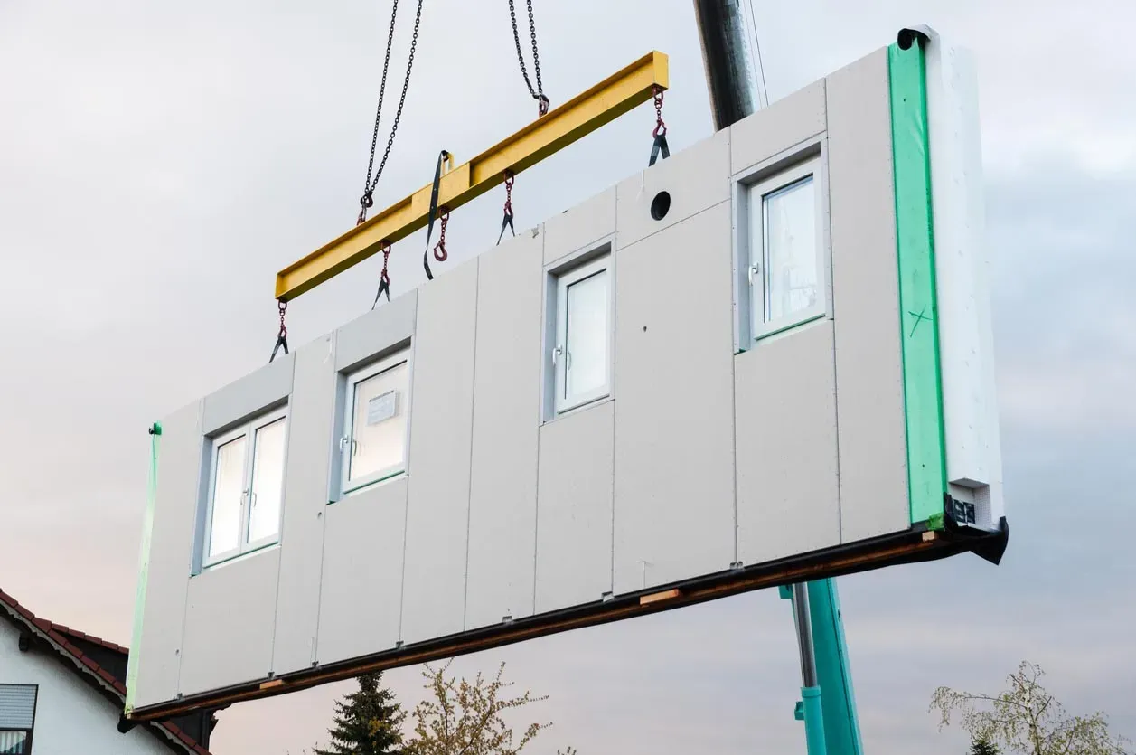 A crane lifts a large pre-fabricated wall panel, complete with three windows, for modular construction.