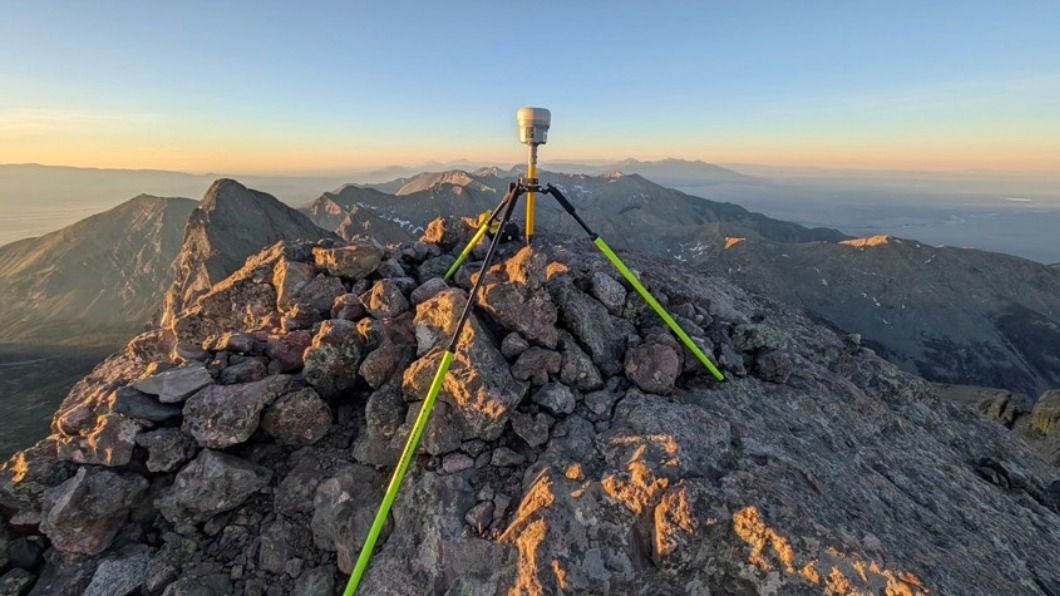 A GPS surveying instrument on a tripod sits atop a rocky mountain summit at sunrise with mountain ranges beyond.