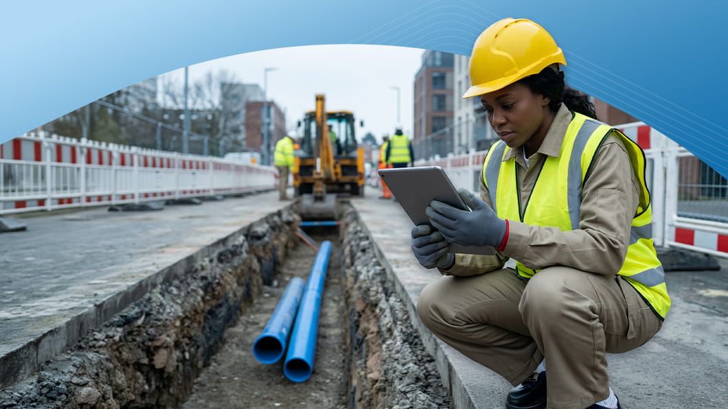 A construction inspector wearing a hard hat and safety vest crouches near an open trench with utility pipes, using a tablet.