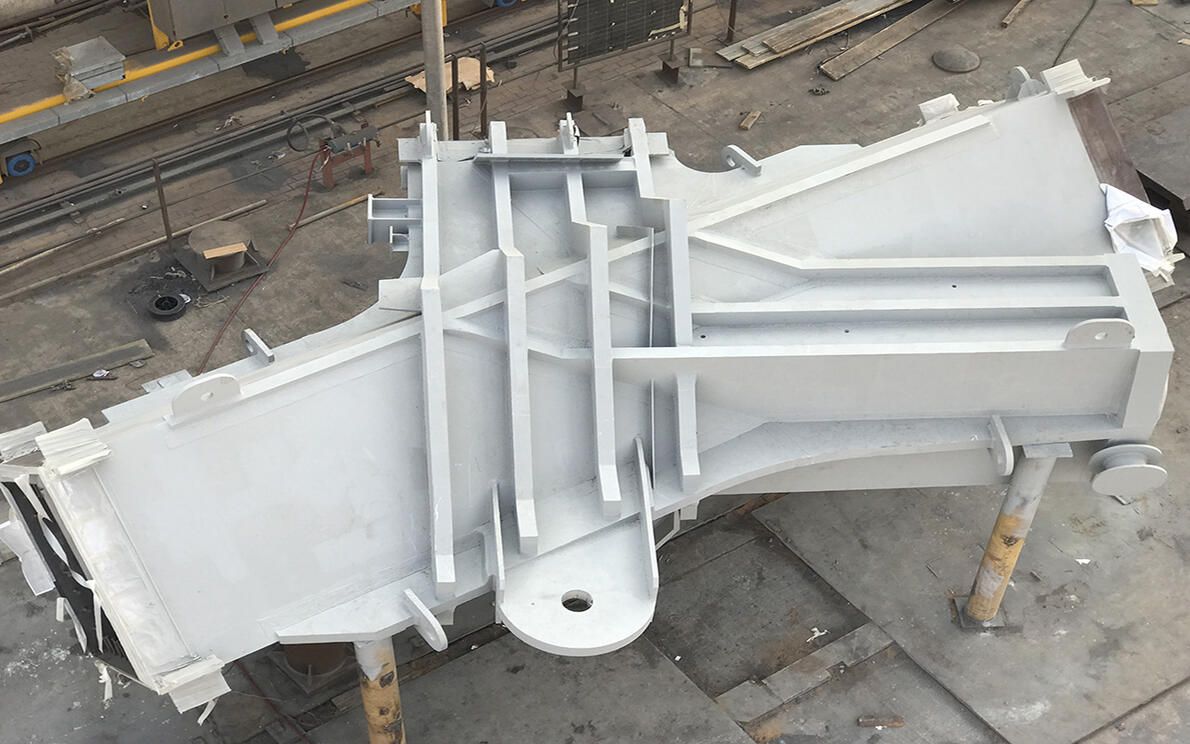 A large, gray, metal component with complex structural supports is shown from an elevated perspective in a workshop.