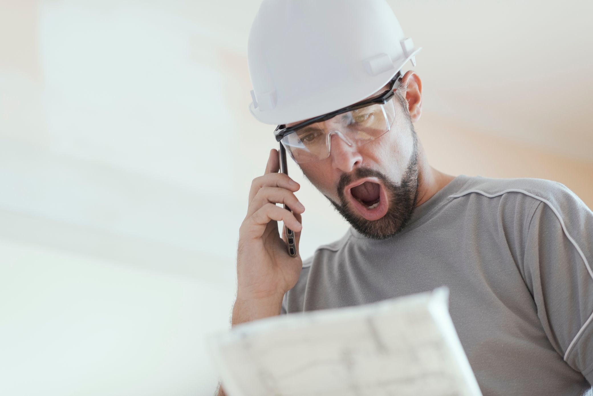 A construction worker in a hard hat and safety glasses looks shocked while talking on cell phone and holding a blueprint.