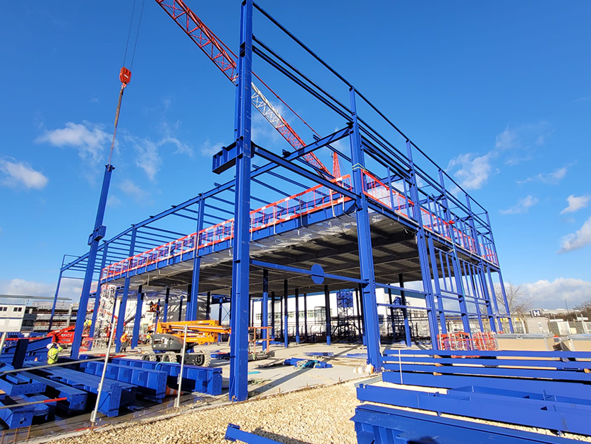 A large steel building frame, painted blue, is under construction on a sunny day with a crane overhead.