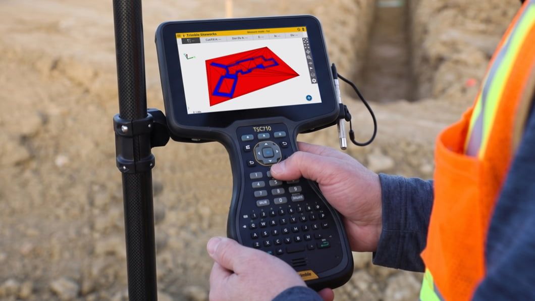 Worker in orange vest using a Trimble TSC710 data collector displaying a 3D site map at a construction site.