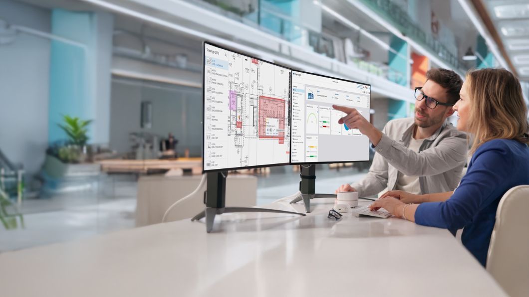 Two colleagues review architectural blueprints and project analytics on dual desktop monitors.