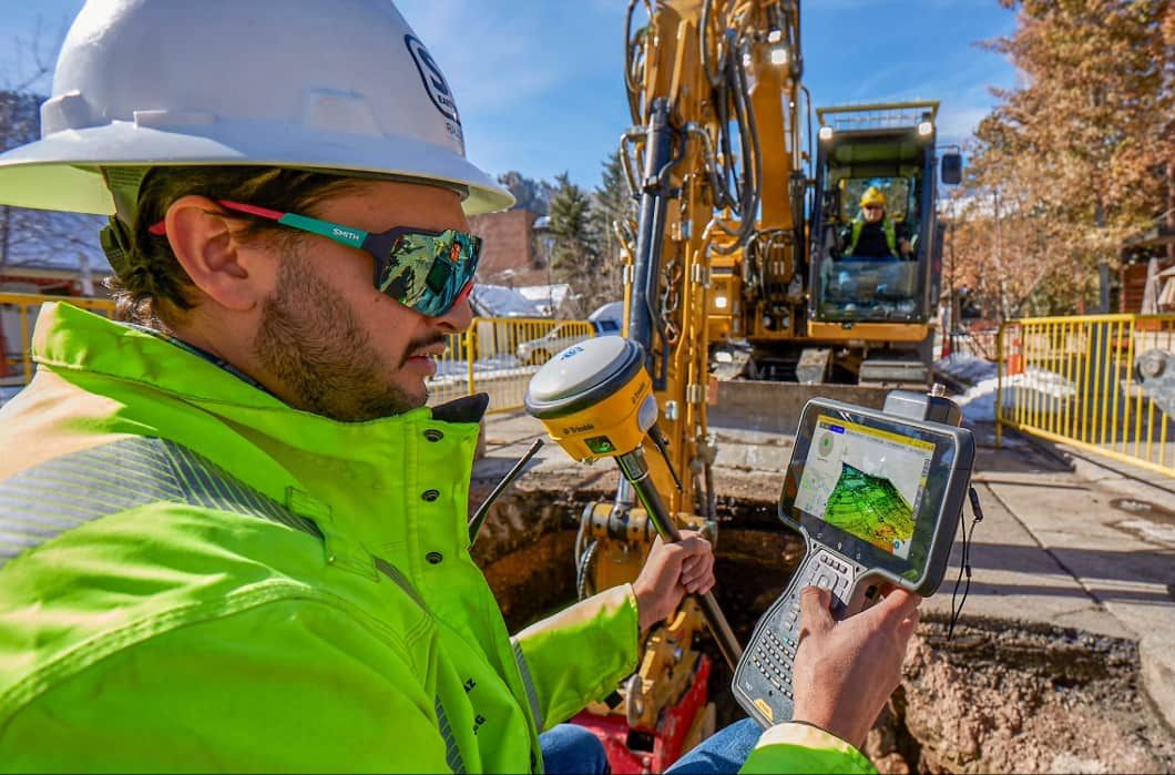 Worker in hard hat and vest uses Trimble GPS and tablet with a digital map, an excavator nearby.