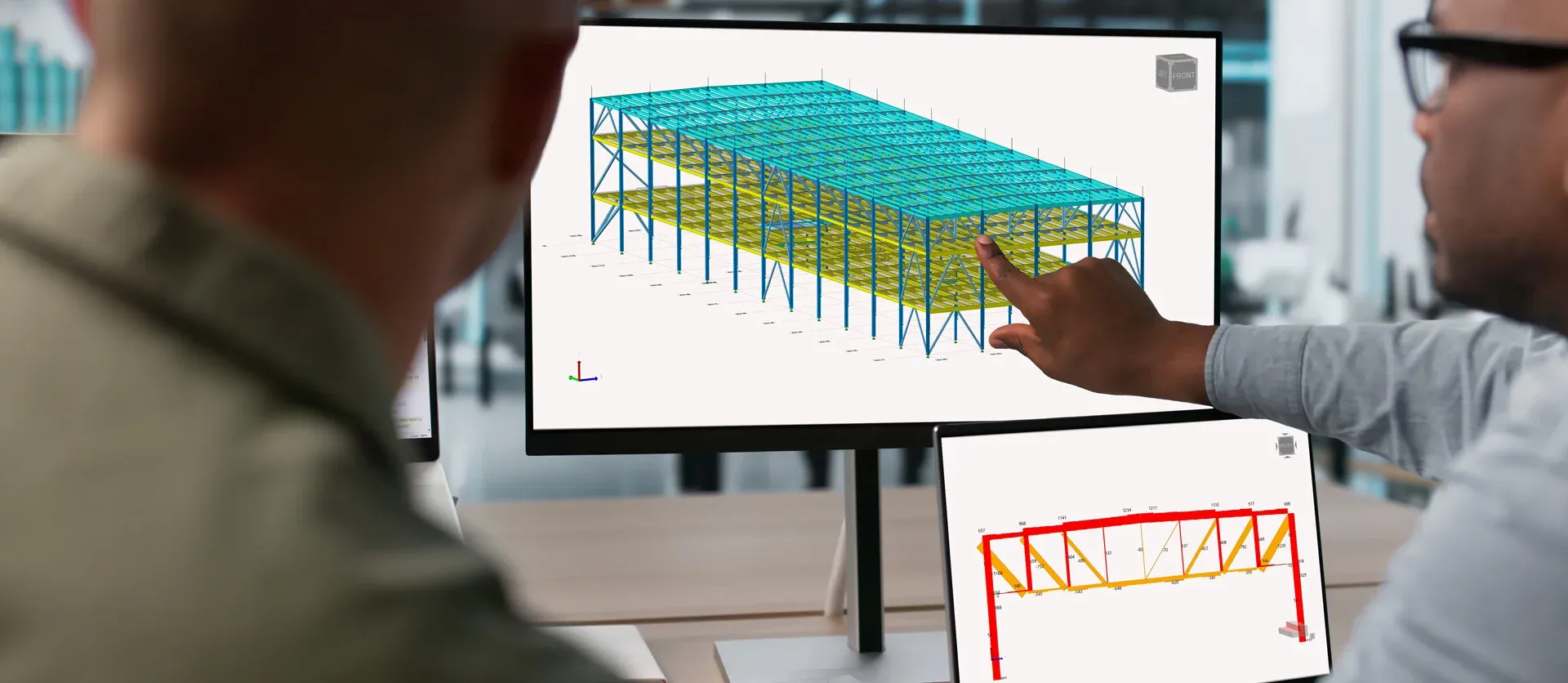 Over-the-shoulder view of two professionals collaborating in front of dual monitors; one person points to a turquoise and yellow 3D structural steel building model on the main screen while a secondary screen displays a red detailed cross-section.