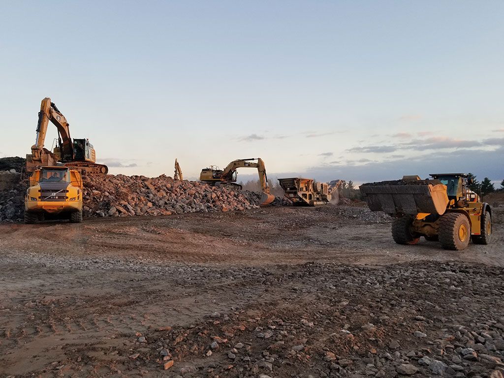Construction vehicles including excavators and a loader operate at a rocky quarry site under a pale sky.