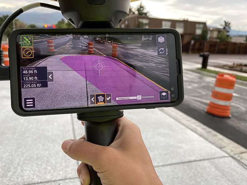 Smartphone displaying augmented reality overlay of construction site measurements with traffic cones in background.