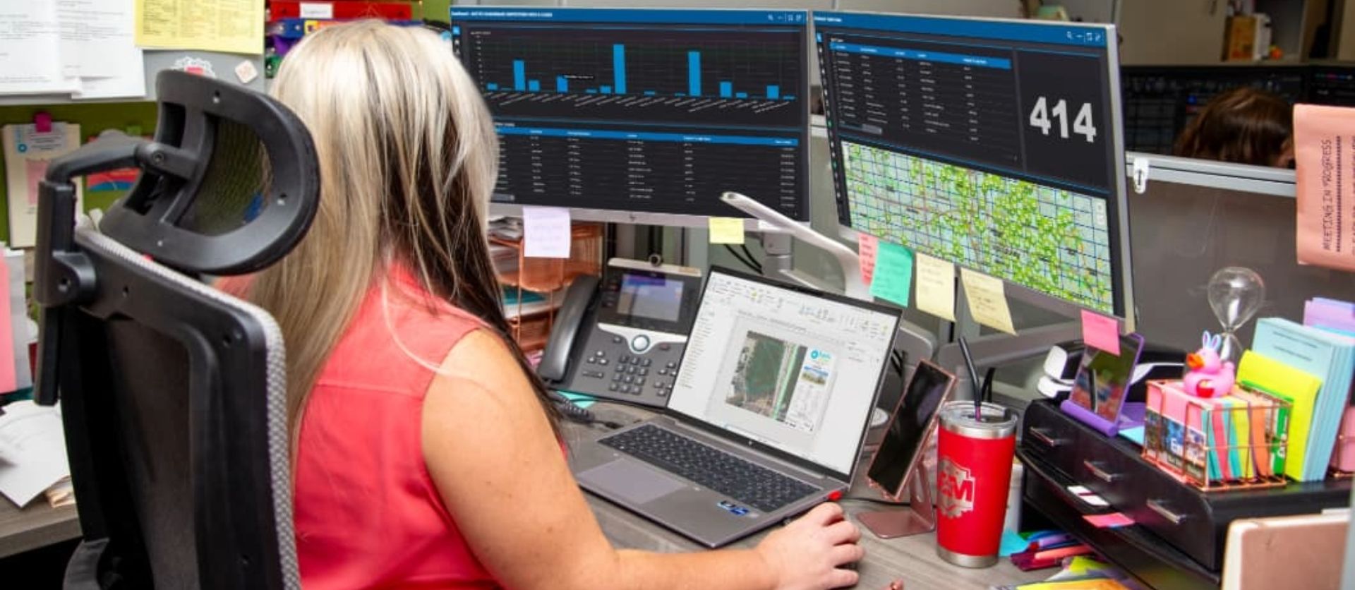 A woman in a cubicle working at a cluttered desk with a laptop and two monitors displaying data dashboards.