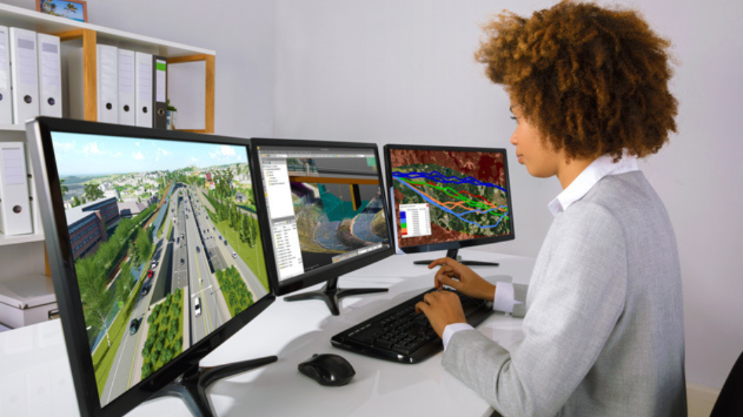 A person at a desk typing in front of three monitors displaying roadway images and data via Trimble Unity software