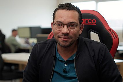 Eine Person in schwarzer Jacke und blauem Hemd sitzt in einem roten Gaming-Stuhl in einem Büroraum.