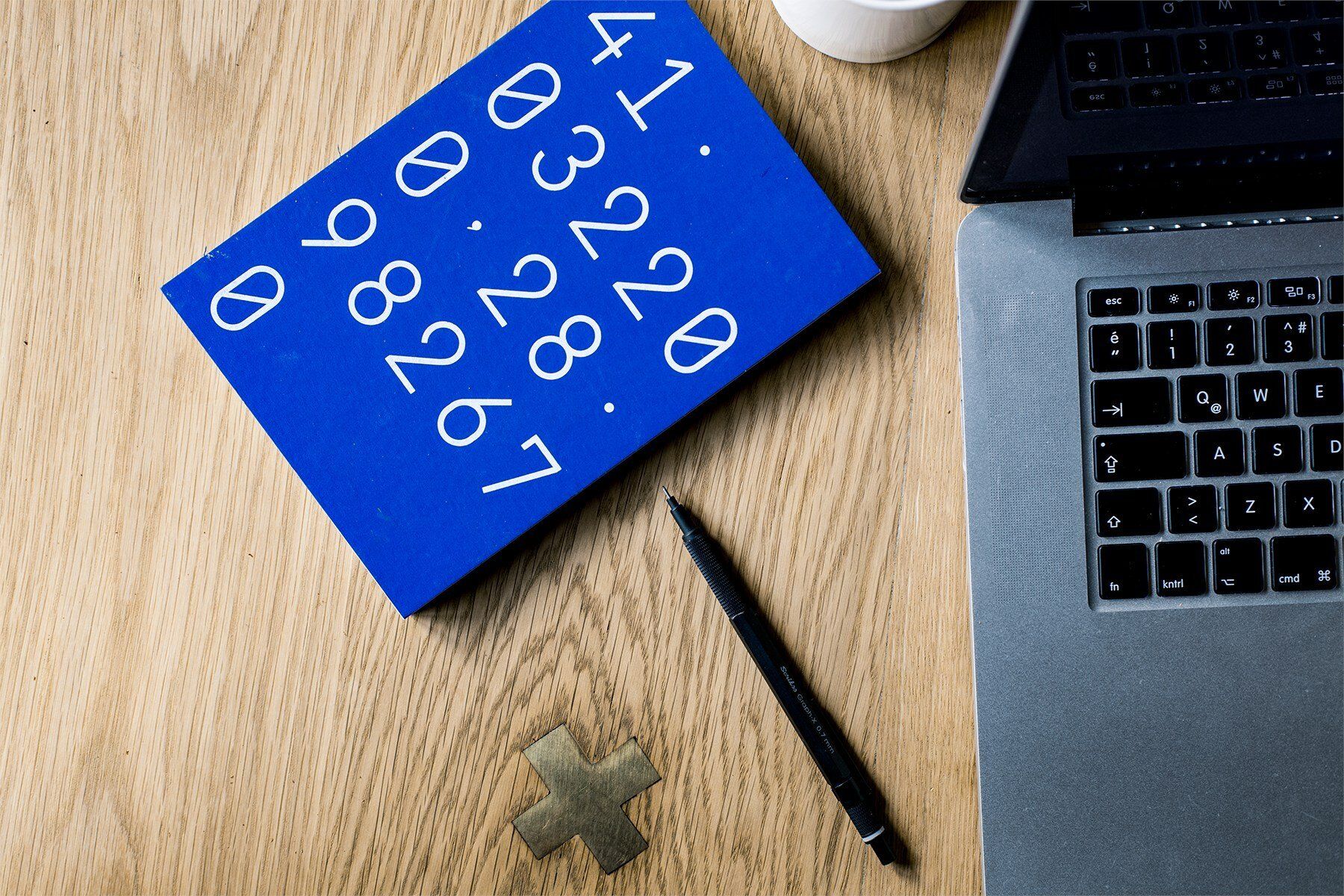 A blue book with white numbers and symbols is next to a laptop, pen, and a small golden cross on a wooden desk.