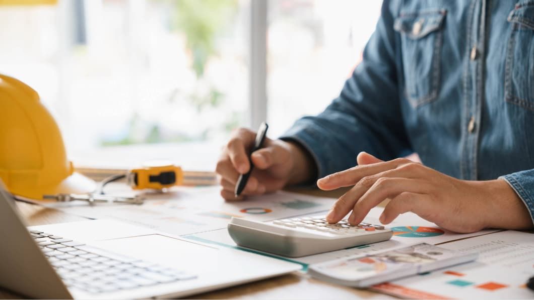 Close-up of a person using a calculator over blueprints and financial charts with a yellow hard hat nearby.