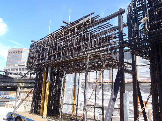 A large, complex cage of rebar, ready for concrete, is shown on a construction site with a modern building in the background.