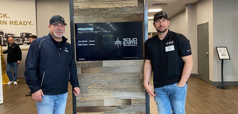 Two men stand by a welcome sign for Solid Earth, showing the owners Dan and Matt Peters.