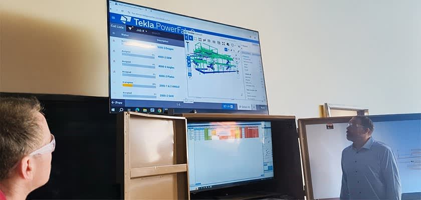 Two men look at a computer screen displaying Tekla PowerFab software with 3D structural modeling.