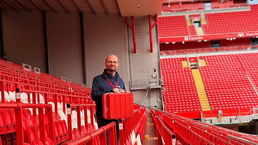 Bald man with Makita lanyard holds red rugged case (survey equipment) in Anfield Stadium. Red seats, Expedia, AXA branding visible.