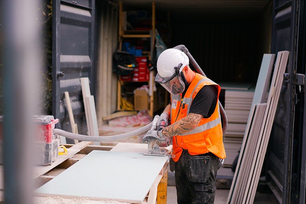 Construction worker in orange safety vest cutting material with power tool at worksite, wearing protective helmet and mask.