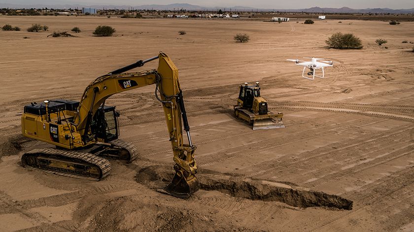 An excavator and bulldozer work on a construction site while a drone flies overhead.