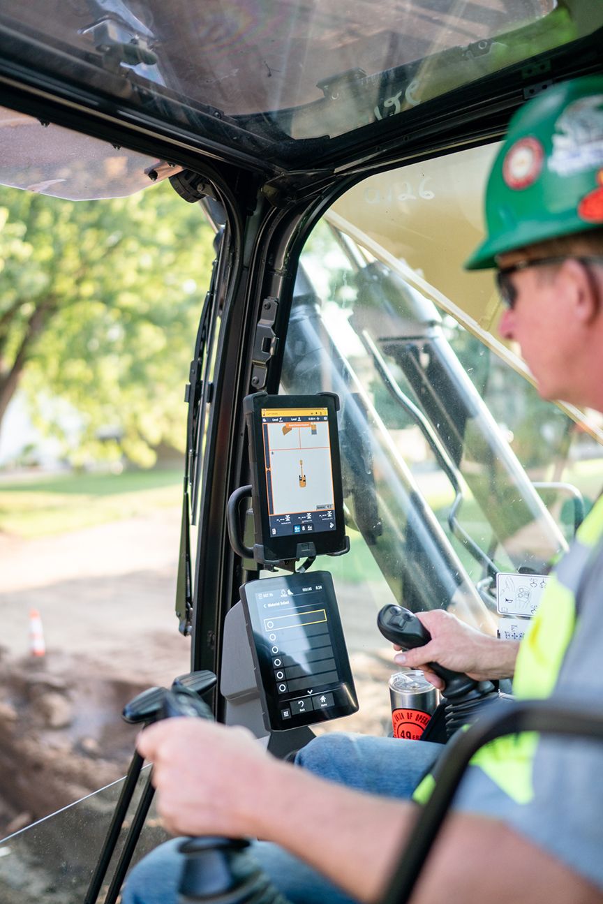 Operator inside an excavator cab using a touchscreen display and joysticks to control machinery.