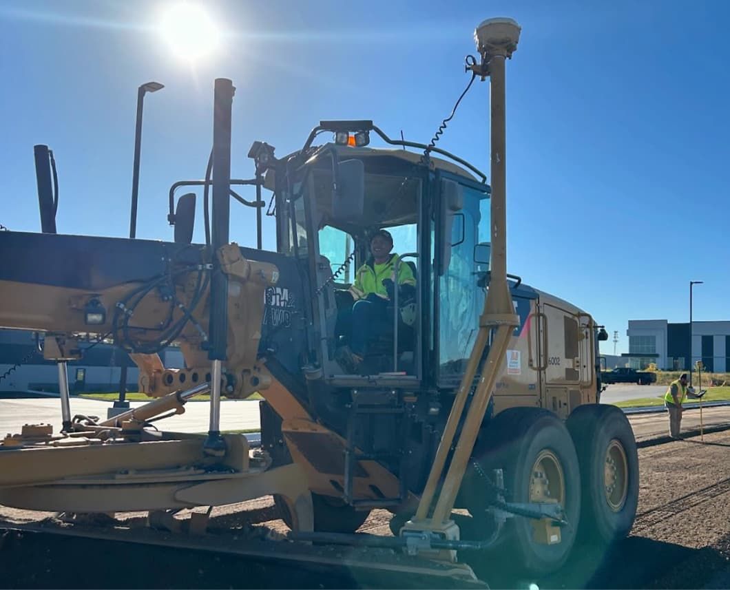 A construction worker in a yellow high-visibility vest operates a yellow grader with a GPS system on a construction site.