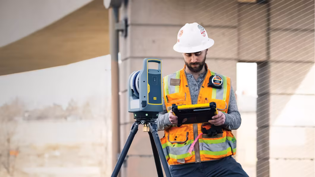 A construction worker in a hard hat and safety vest uses a tablet next to a Trimble X9 laser scanner.
