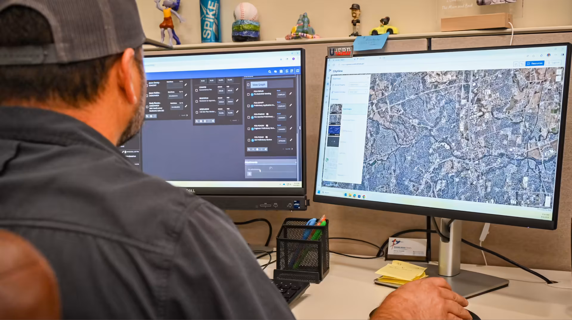 Person wearing a cap works at a desk with two monitors; one shows a control panel interface, and the other displays a detailed satellite map.
