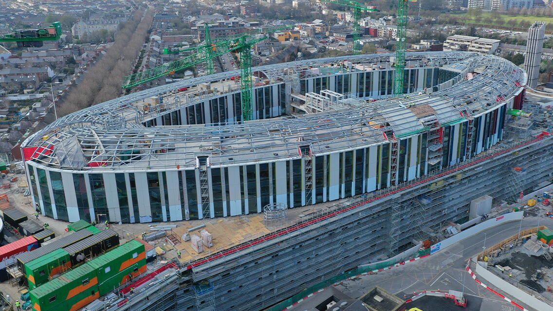 Aerial view of a large, oval-shaped stadium under construction, with the steel framework of the roof clearly visible.