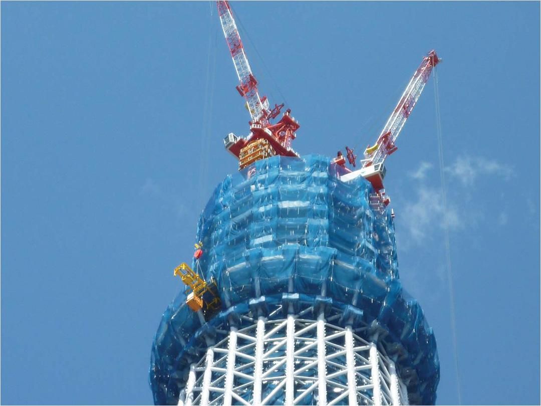 Close-up of the Tokyo Skytree under construction, with cranes at the top and blue netting covering the structure.