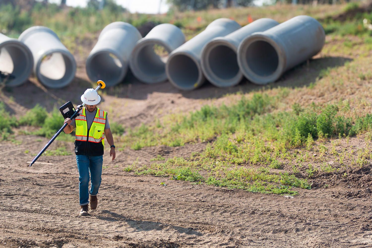 A surveyor walks on a construction site carrying surveying equipment, with concrete pipes in the background.