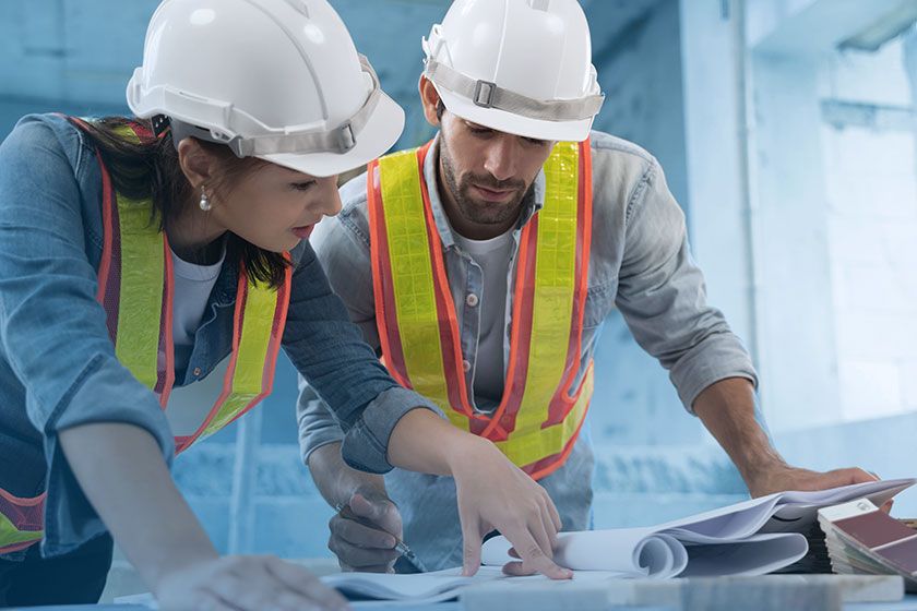 Two construction workers, a woman and a man, wearing hard hats and high-visibility vests, collaborate over blueprints.