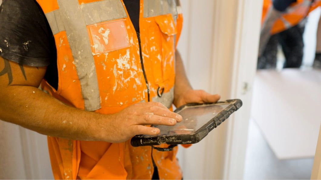 Construction worker in orange safety vest using a tablet device on a job site, with dust-covered hands.
