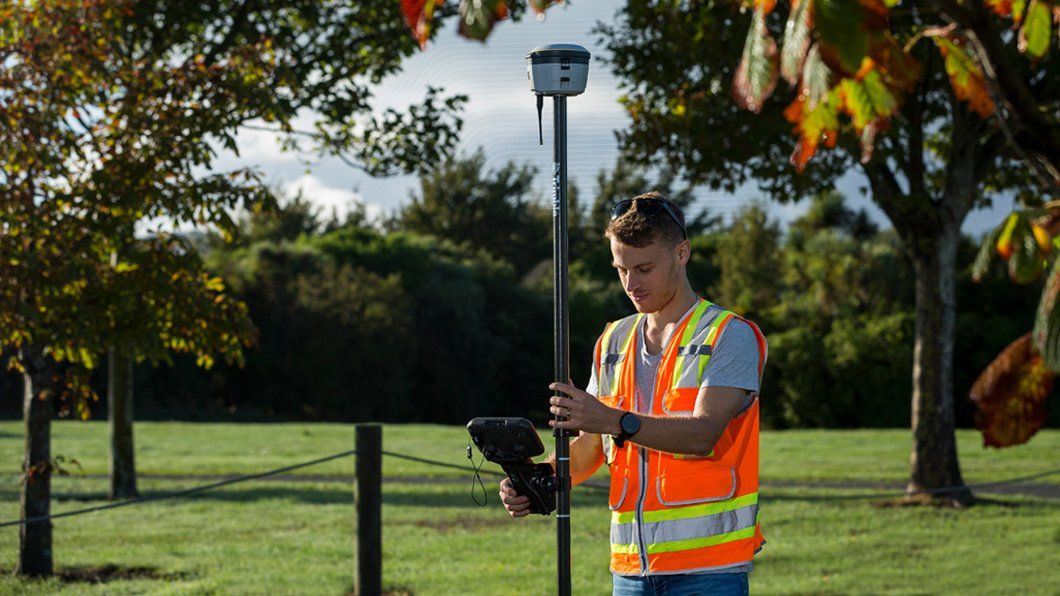 Surveyor in hi-vis vest uses Trimble R10 GNSS receiver on range pole with TSC7 controller for precise site data collection.