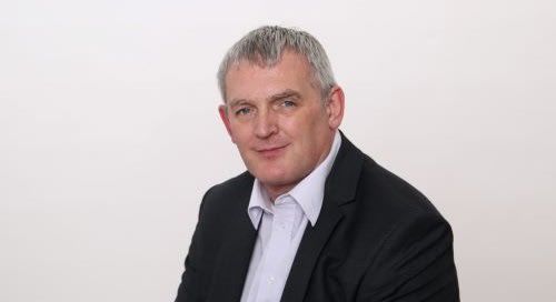 A headshot of a middle-aged man with graying hair, wearing a black suit jacket over a light purple shirt.